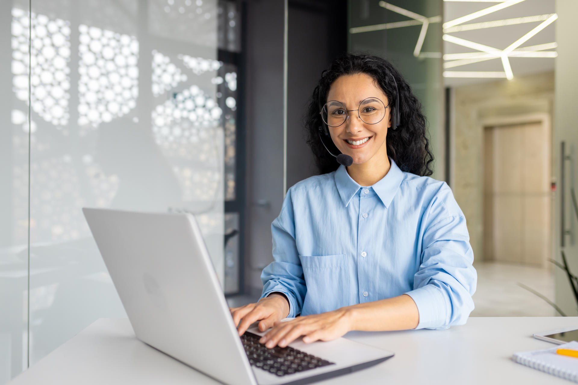 Portrait of successful woman helpline worker, using headset phone for video call, smiling and looking at camera, indian woman happy with dream job, inside office with laptop.
